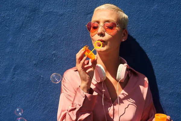 Never let anyone burst your bubble. an attractive young woman standing against a blue wall alone and blowing bubbles while outdoors