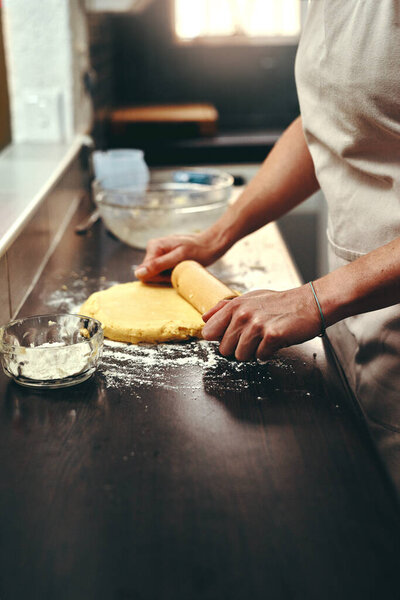 This will be definitely baked to perfection. an unrecognizable woman flattening dough with a rolling pin while baking inside her kitchen at home