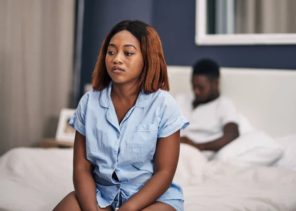 She doesnt know what to do. an attractive young woman looking depressed while sitting on the edge of her bed with her husband in the background