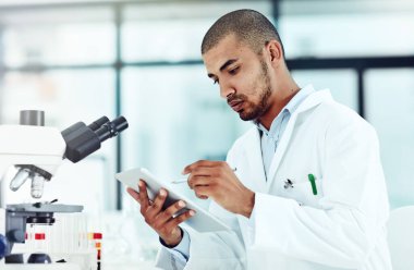 Serious male scientist working on a tablet reviewing an online phd publication in a lab. Laboratory worker updating health data for a science journal. Medical professional document clinical trial.