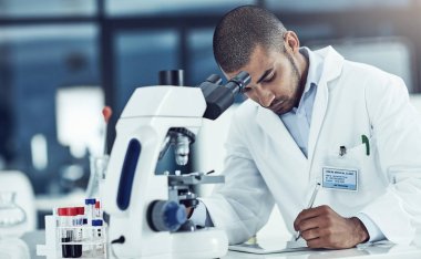 Focused male lab technician using digital tablet working on medical development and innovation project with microscope, taking notes. Doctor diagnosing test tube blood sample with science equipment