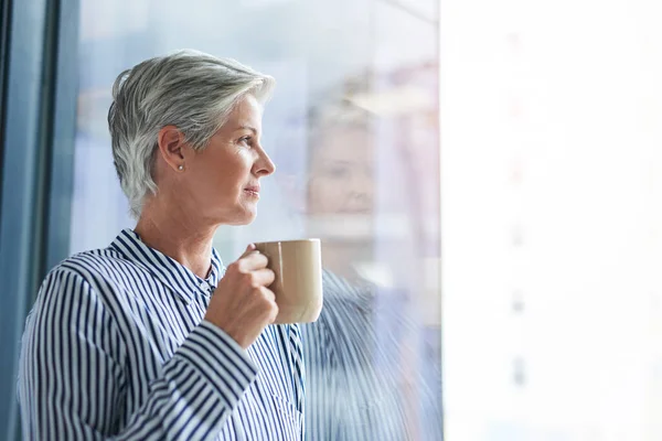 You grow wiser in thought as the years go by. an attractive mature businesswoman drinking coffee while looking out of her office window