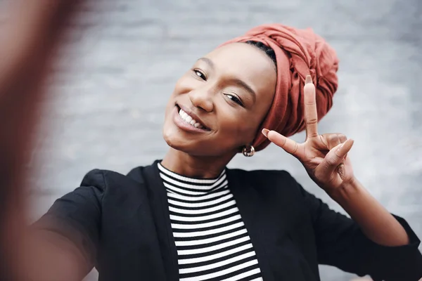 Spread peace. Cropped portrait of an attractive young businesswoman taking selfies while gesturing the peace sign against a grey brick wall outside