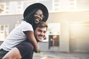 Young playful interracial couple playing, bonding and hugging while standing in the city together on weekend. Happy husband giving wife a piggyback ride, enjoying a day in the urban town or traveling.
