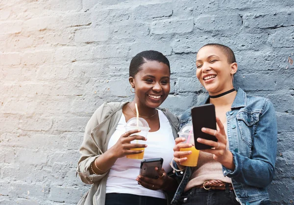 Sometimes you have to see it to believe it. two young women standing beside a building smiling and reading through text messages while holding their cool drinks