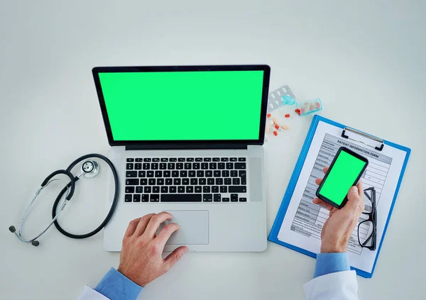 Technology helps keep track of it all. High angle shot of an unrecognizable doctor using a laptop and cellphone with a green screen in his office