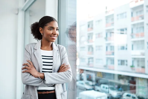 Young professional businesswoman, smiling while looking out of window of her modern office, thinking in her leadership role. Happy female manager standing with the vision of ambitious motivation.