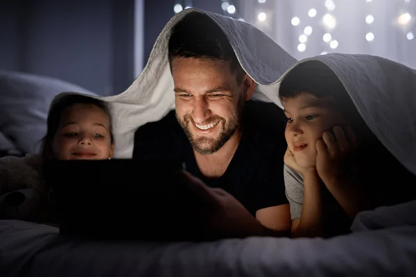 Whether its day or night, they stay entertained. a father and his two little children using a digital tablet together in bed at night