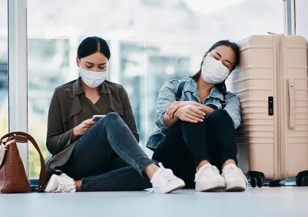 Young women in quarantine after travel, wearing safety mask to protect from covid and sitting in an airport traveling together during pandemic. Tired female friends leaving for getaway vacation.