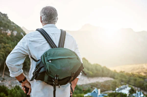 . Rear view of exploring, active and adventure senior man standing with a backpack after a hike, enjoying the landscape and forest nature. Male hiker looking at nature environment sunrise view