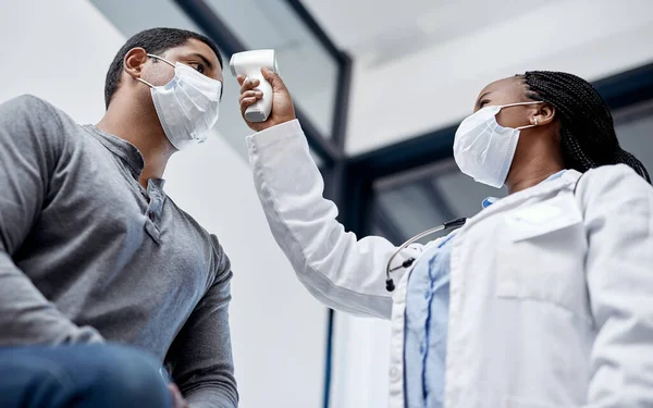 Female doctor consulting a male covid patient, taking his temperature with infrared thermometer in hospital room or clinic. Man wearing mask and health care professional pointing medical equipment