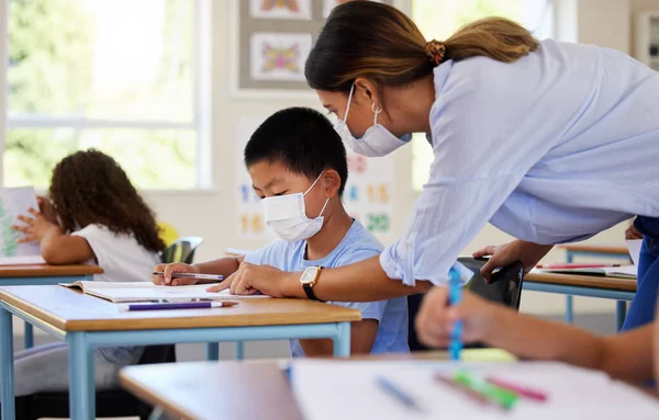 Education, covid and learning with face mask on boy doing school work in classroom, teacher helping student while writing in class. Elementary child wearing protection to stop the spread of a virus.