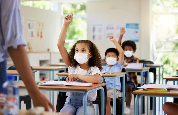 Covid, education and learning of eager, smart and clever children raising hands and wearing masks in a classroom. Teacher or educator asking questions for knowledge test at kids school in a pandemic