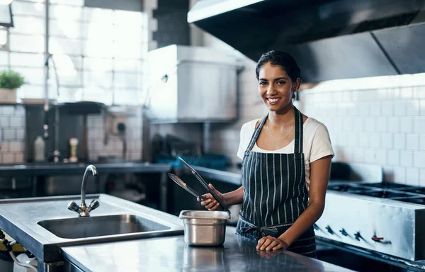 Cooking, making food and working as a chef in a commercial kitchen with tongs and industrial equipment. Portrait of a female cook preparing a meal for lunch, dinner or supper in a restaurant or cafe.
