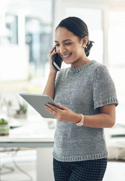 Professional smiling businesswoman talking on phone call with client in modern office. Young design employee working on appointment booking on tablet standing in casual startup company.