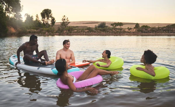 Diverse friend group floating on lake water, having fun in nature and bonding on a vacation in countryside sunset with inflatable rings. Men and women laughing, smiling and looking relaxed on holiday.