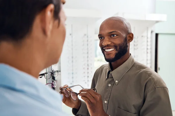 Shopping for glasses at an optician retail store with a smiling man trying to search for a pair. Optometrist and customer service employee selling a happy buying client new and modern spectacles.