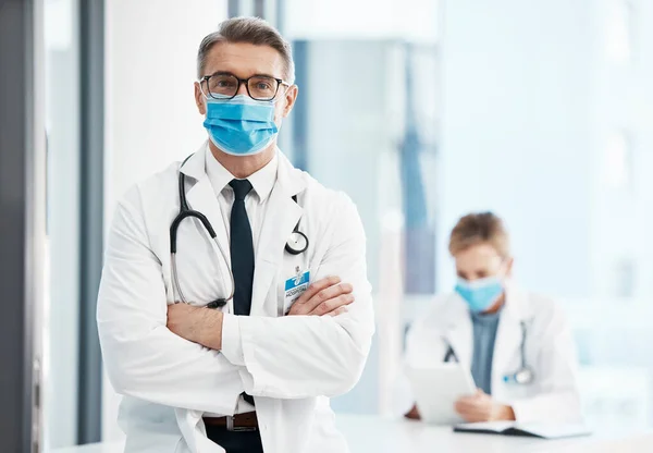 Doctor, physician or healthcare professional with covid face mask in a hospital for medical health insurance background. Innovation, leadership and excellence male gp portrait with his arms crossed.
