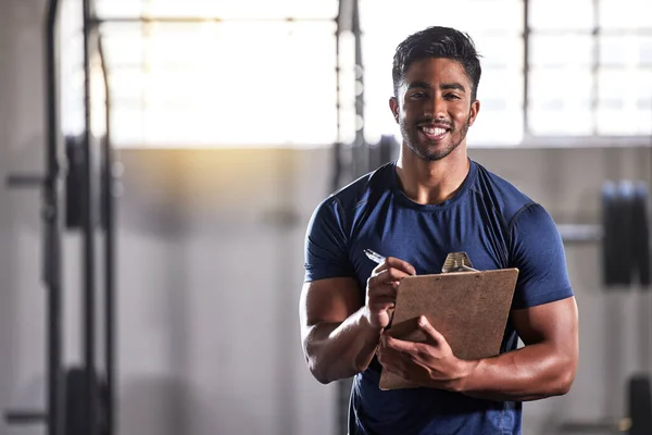 Gym, workout and personal trainer with clipboard consulting at a training sports in gym. Portrait of muscular, active and smiling fitness coach writing on health, wellness and exercise with flare.