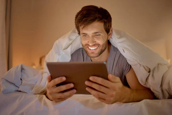 Happy man relaxing in bed with digital tablet watch, movie, series or online social media videos on an app. Browsing the internet news with 5g technology on wireless device in the bedroom at home.