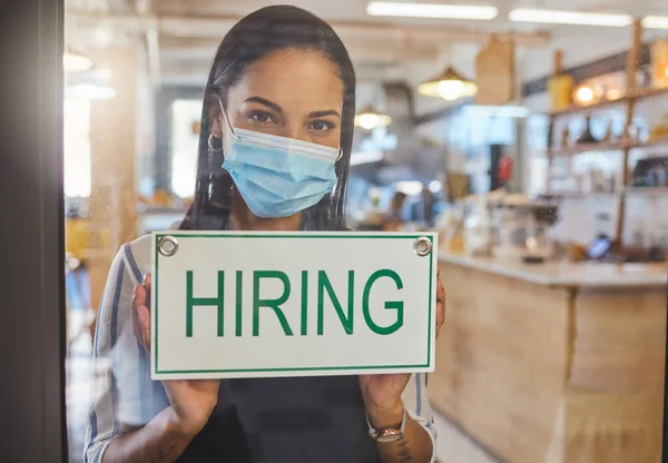 Business woman advertising a hiring sign in her startup coffee shop door. Entrepreneur, business owner or manager wearing a mask searching for employee or workers in a cafe during covid pandemic.