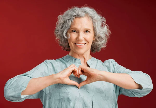 Love, heart and health retired woman with loving emoji sign, icon or symbol showing care or affection on red valentines day studio background. Grey, senior or pensioner with a trendy emoticon pose.