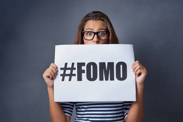 If it happened online it must be real. Studio shot of an attractive young woman holding a sign with FOMO printed on it against a gray background