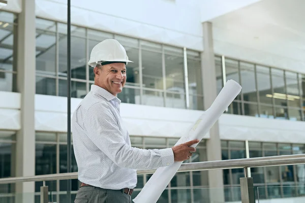 You can count on him to get the job done. Portrait of a cheerful professional male architect looking at the camera while holding blueprints inside a building