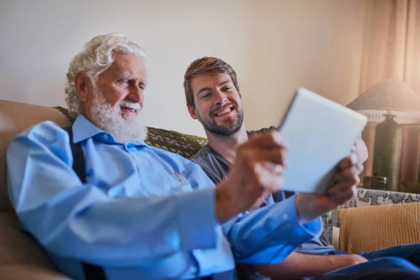 Let your grandpa show you a thing or two. a young man showing his elderly grandfather how to use a tablet while sitting on the couch at home