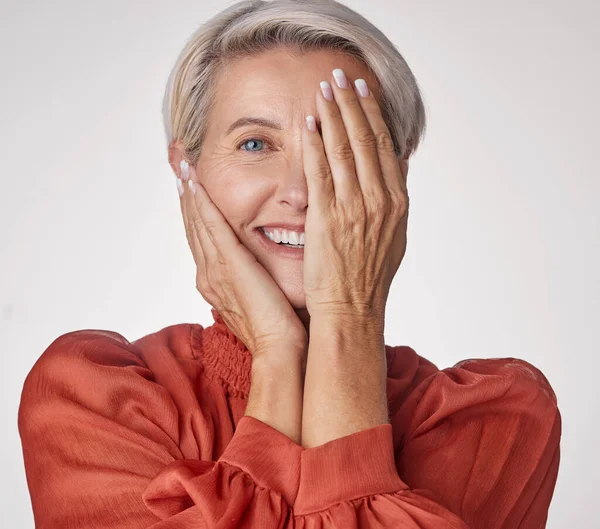 Eye, half and smile of a senior woman isolated against a grey background in a studio. Portrait of an elderly model lady in beauty with hands over her face in wellness, health and teeth