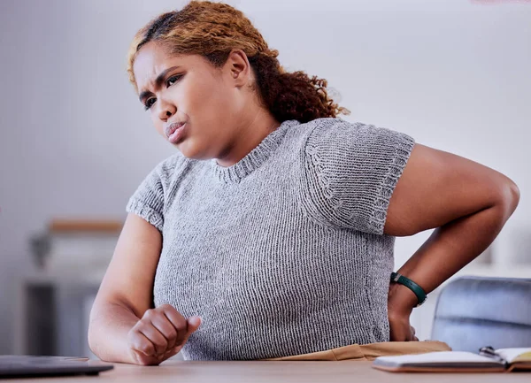 Corporate employee suffering from back pain while working at a desk in an office, uncomfortable and concerned. Young professional experience discomfort from an injury, bad posture or hurt muscle.