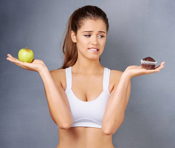 What about a bite of each then. Studio shot of a young woman choosing between healthy and unhealthy foods against a grey background