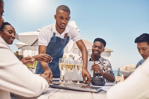 Beach restaurant, waiter and champagne celebration with friends. Hospitality, fine dining and a relaxing outdoor meal with people. Summer holiday or lunch break outside with view of the ocean