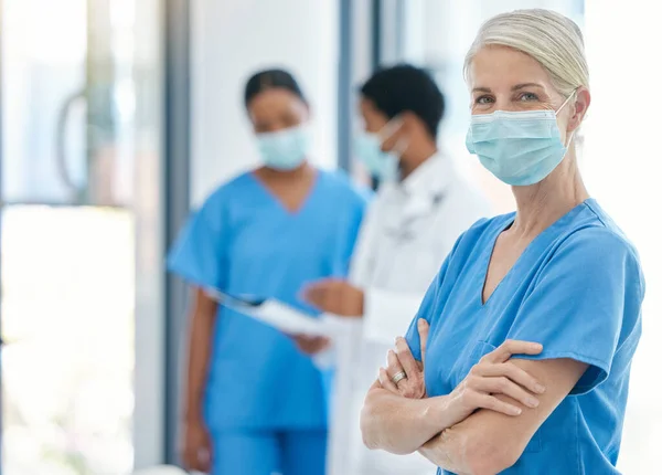 Success, healthcare and leadership, woman doctor in surgical mask and scrubs, standing arms crossed with coworkers talking in background. Proud, professional and motivated to fight covid at hospital