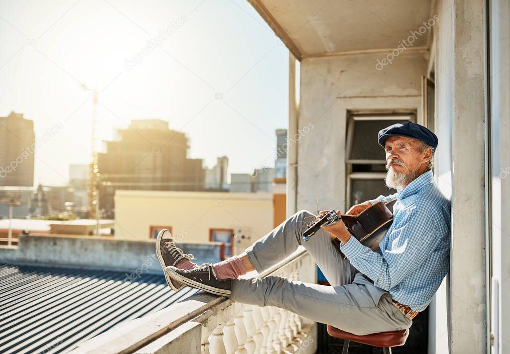 Working on his latest song. a peaceful senior man sitting on his balcony while playing acoustic guitar with the city in the background