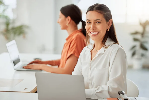 Small business owner or startup entrepreneur working on a laptop in the office with a colleague in the background. Typing an email or planning ideas at her desk in a modern and creative company.