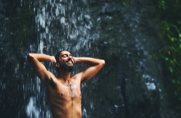 Nature is bliss. a young man standing under a waterfall being completely soaked in water but loving every moment of it