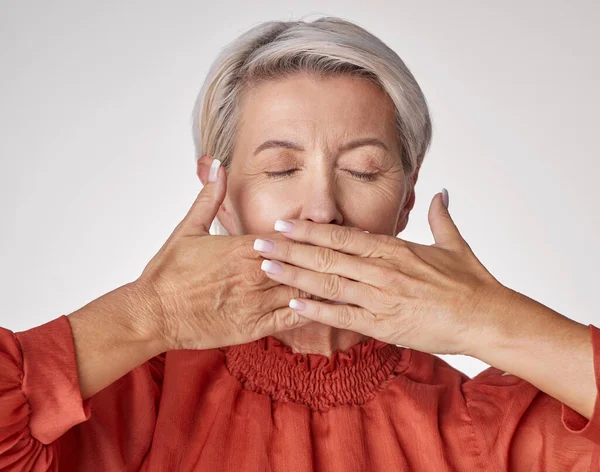 Senior, hands and mouth of an elderly woman covering her teeth or lips with hand against a grey studio background. Mature and aging woman keeping secrets, not talking and hiding the truth or gossip