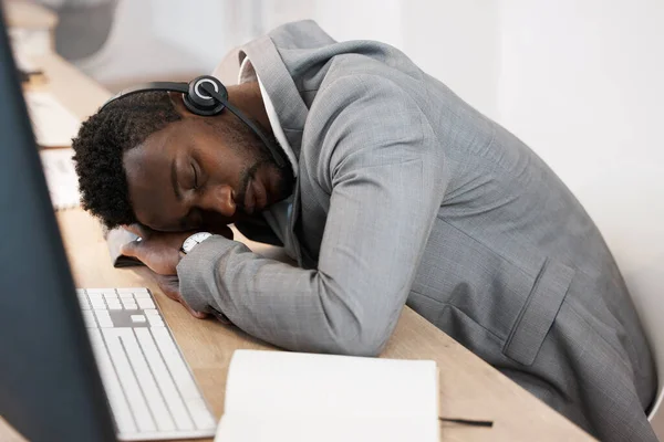 Tired CRM call center agent sleeping on computer desk at work office with headset on. Exhausted young business man taking a nap on table. African American male feeling burnout resting in workplace