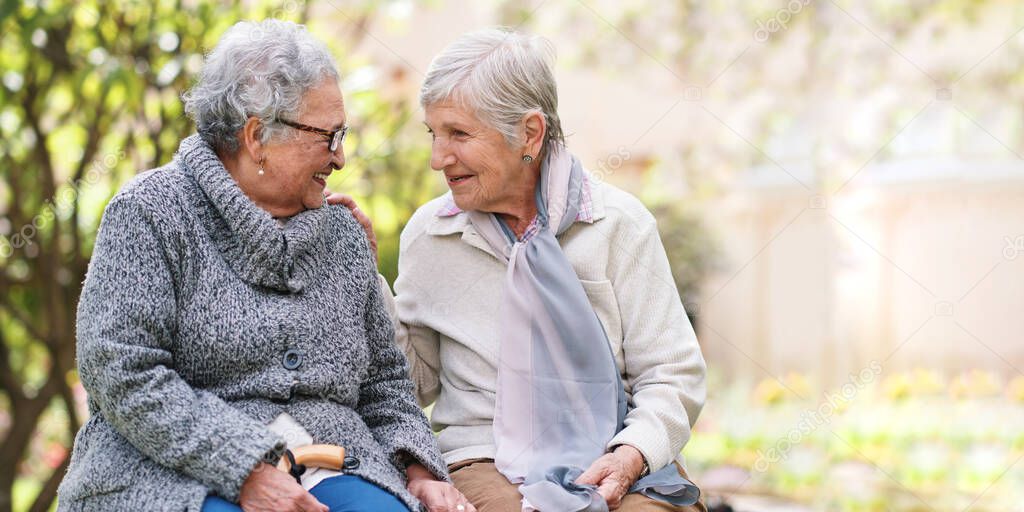 Two elderly women sitting on bench in park smiling happy life long friends enjoying retirement.