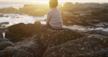 Ocean, freedom and thinking child by the rocks outdoors sitting by the water with a relaxing sunrise. Nature, travel and young boy on summer vacation sitting by sea or rock beach watching the waves