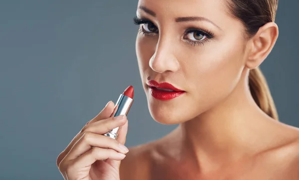 Vamp up your makeup routine with red lipstick. Studio portrait of a beautiful young woman putting on red lipstick against a grey background