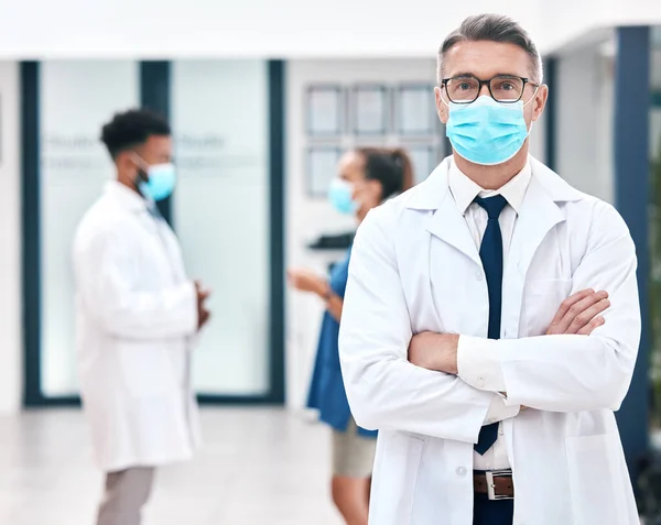 Mask, covid and medicine with a man doctor wearing a mask in a hospital for safety, health and wellness. Healthcare, medical and insurance with a male surgeon standing arms crossed in a clinic.