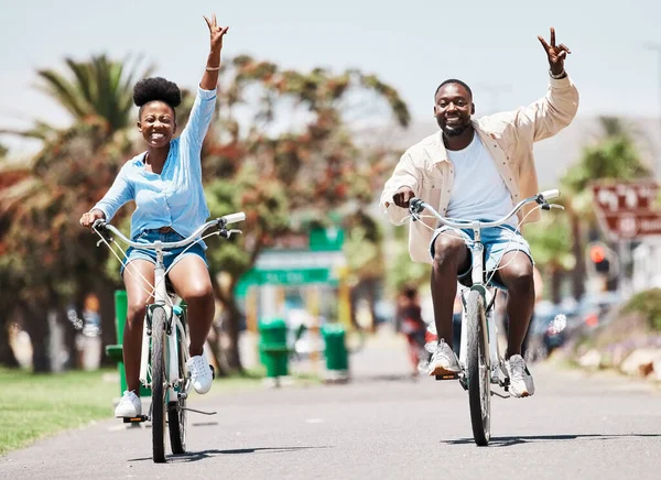 African couple cycling on bike on vacation, peace hand sign on bicycle for sustainable lifestyle in the city and happy on holiday in summer for travel. Eco friendly man and woman in Miami for spring.