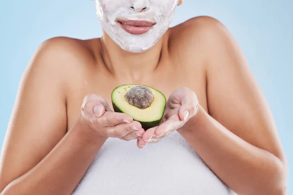Hands, avocado and skincare face mask of a woman in beauty and cosmetic facial on a blue studio background. Female in health, skin and body care holding green fruit for nutrition, food or diet.