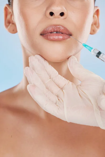 Closeup of a young woman getting lips injection treatment from a cosmetic doctor in a studio. Syringe and needle with botox filler for the mouth by a cosmetology beautician with a blue background