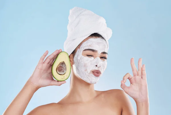 Face mask, avocado and woman with a ok sign with health, wellness and organic lifestyle in studio. Girl doing fresh, clean and natural selfcare routine while holding fruit for nutrition and diet