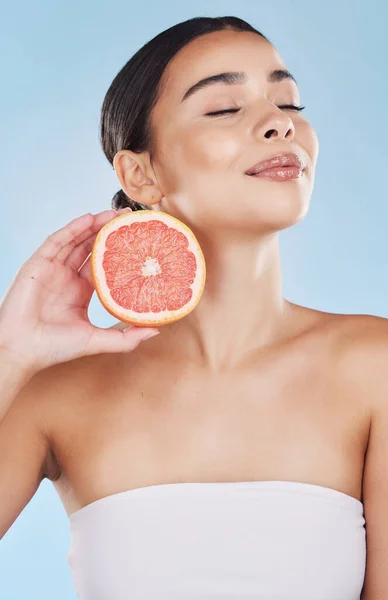 Grapefruit, woman skincare and wellness fruit for face grooming, wellness and diet health on a blue background in studio. Happy, smile and beauty brazilian model with vitamin c food product in hands.