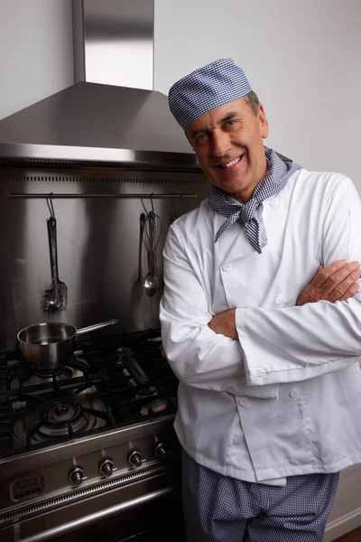 Smiling senior chef with hands folded standing by gas stove. Portrait of a smiling senior chef with hands folded standing by gas stove and vent hood