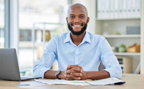 Portrait of a happy African businessman sitting at his desk in his modern corporate office. Professional black manager working or planning a company project with a laptop and paperwork at a table